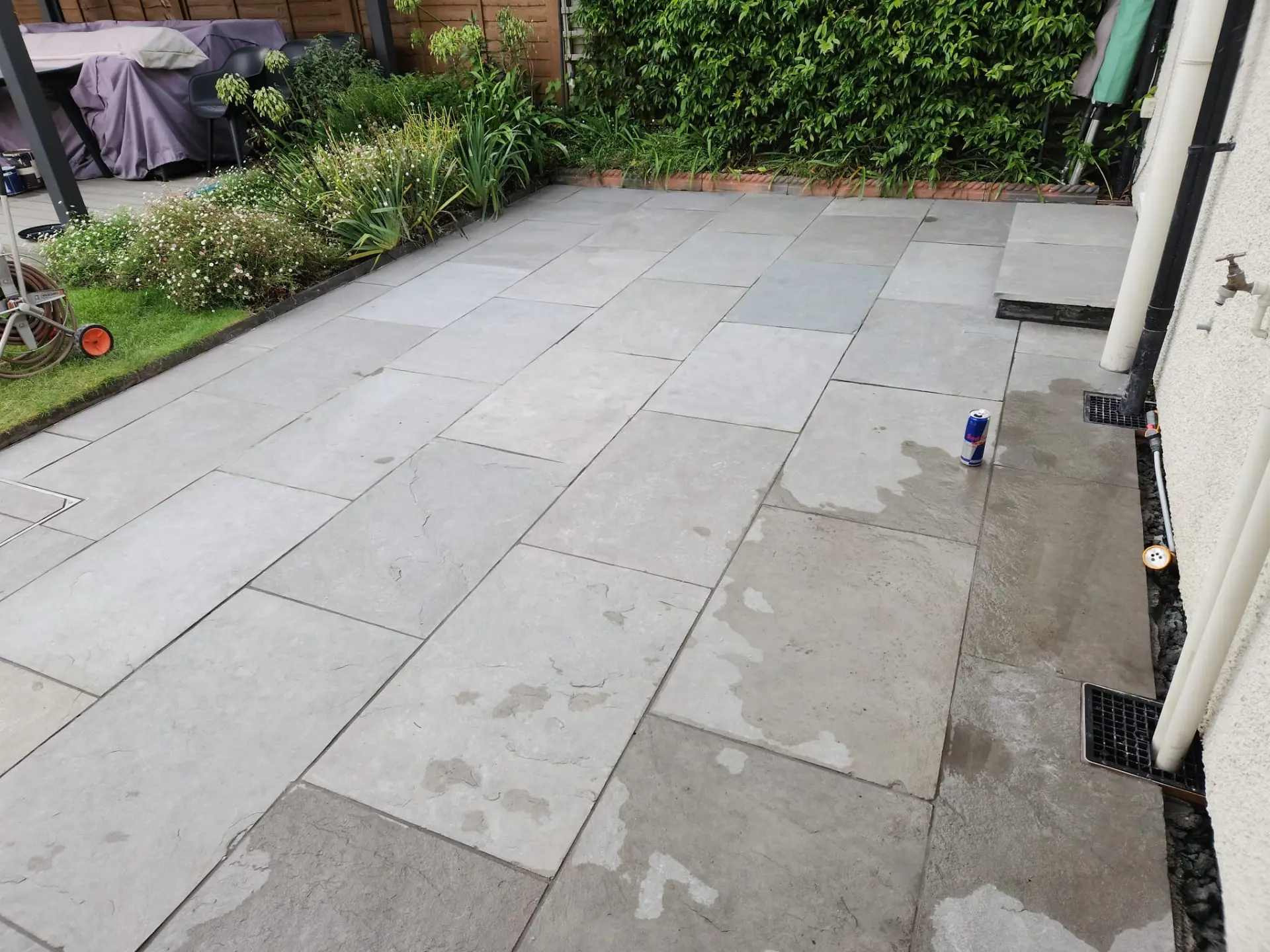 Large grey sandstone patio laid across a rear garden with planted border and drainage channel