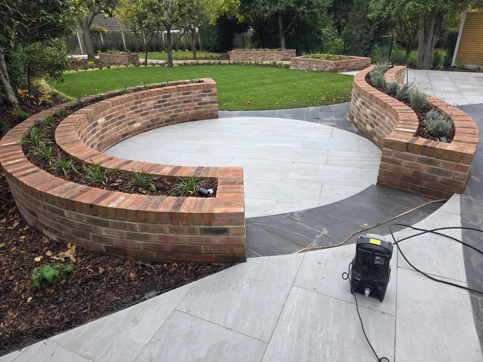 Circular porcelain patio framed by curved red brick retaining walls and planted borders