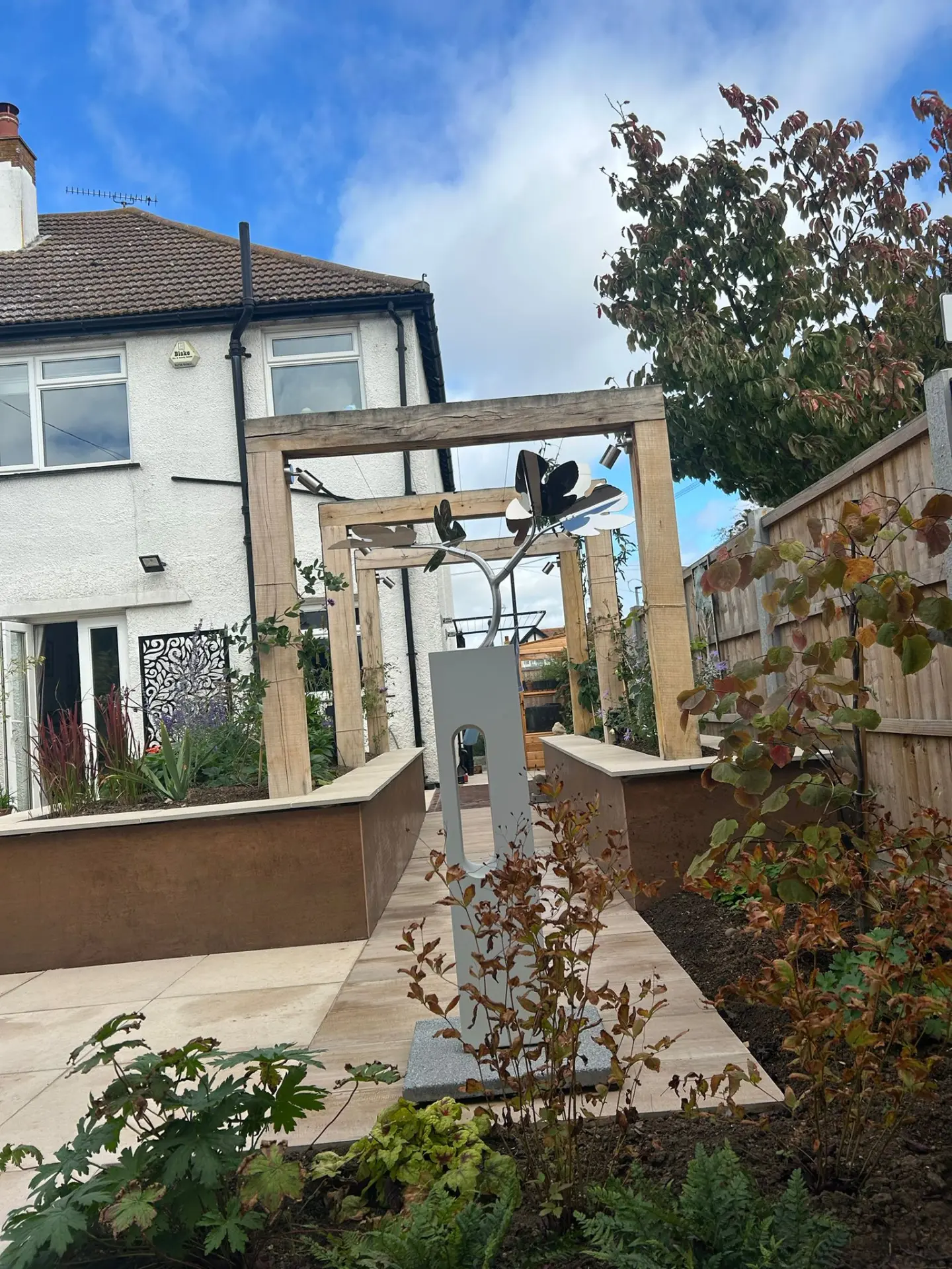 Oak pergola frames over a stone path with corten steel raised beds and a metal garden sculpture